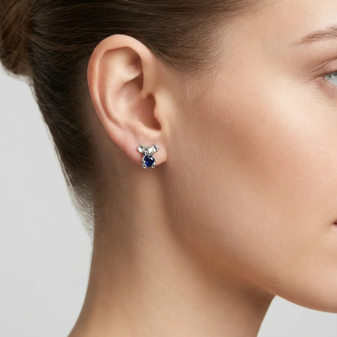 Close-up of a woman wearing a pair of Koala earrings with a heart shape blue CZ on a neutral background
